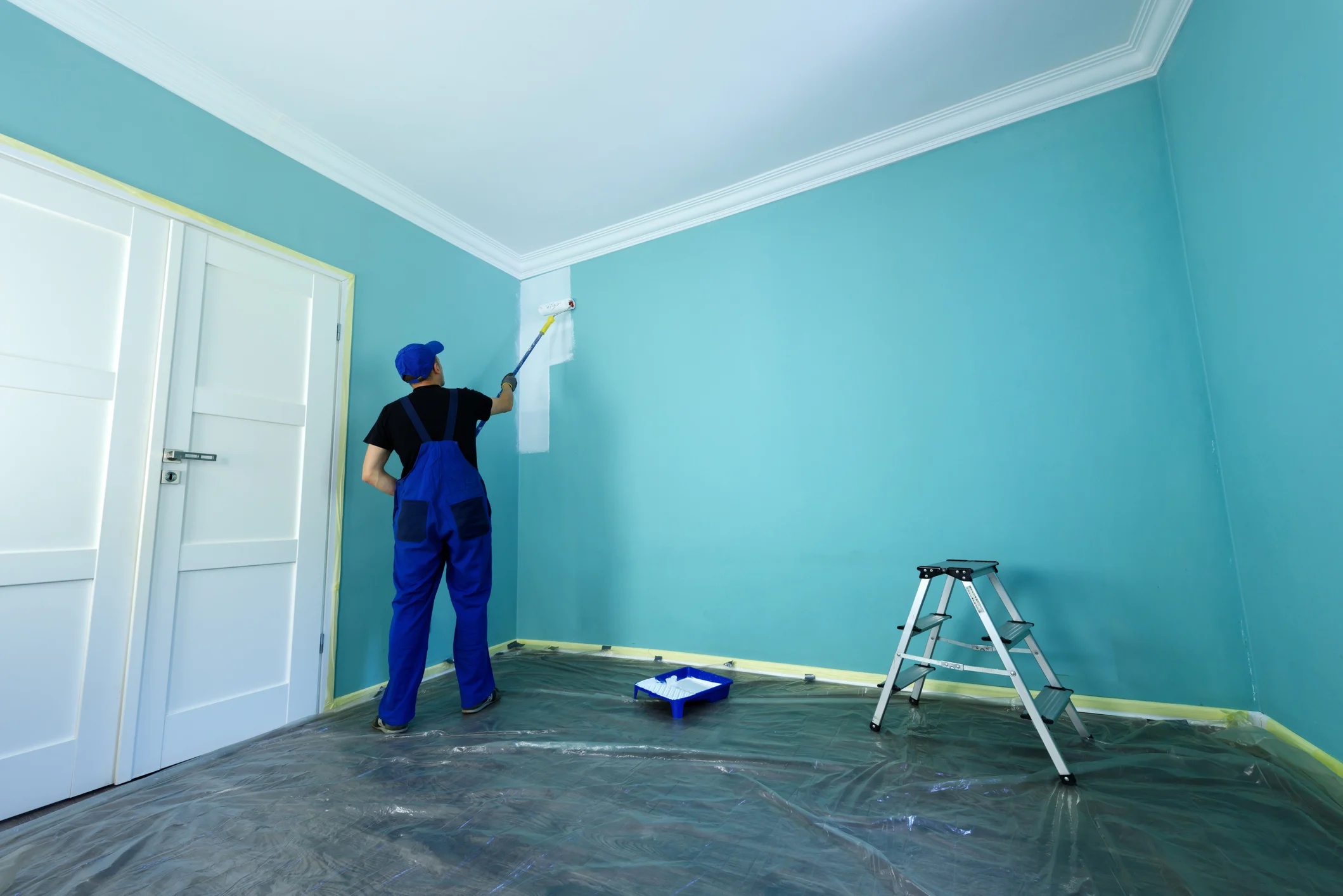 Painter applying a soft blue interior wall colour with a roller in an empty, fully masked room
