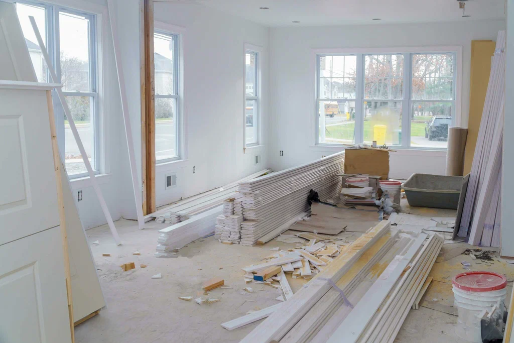 Interior of a bright room under renovation with stacks of white trim, paint buckets, and construction materials