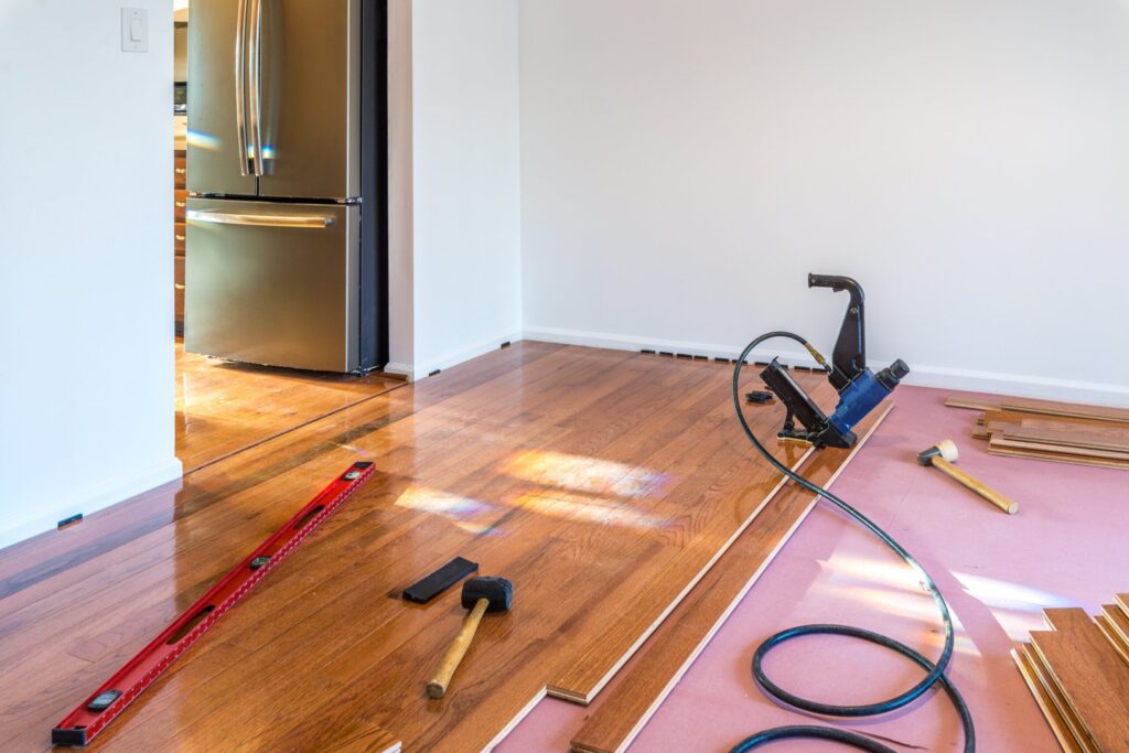 Hardwood floor installation in progress with a pneumatic flooring nailer, rubber mallet, and level on new wood planks